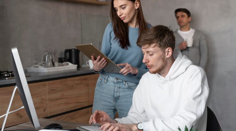 Служители в компания, мъж работи на лаптоп и права жена до него на таблет, трети пие кафе на заден фон